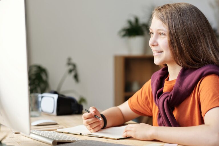 A smiling teenage girl engages in online learning at her home workspace, using modern technology.