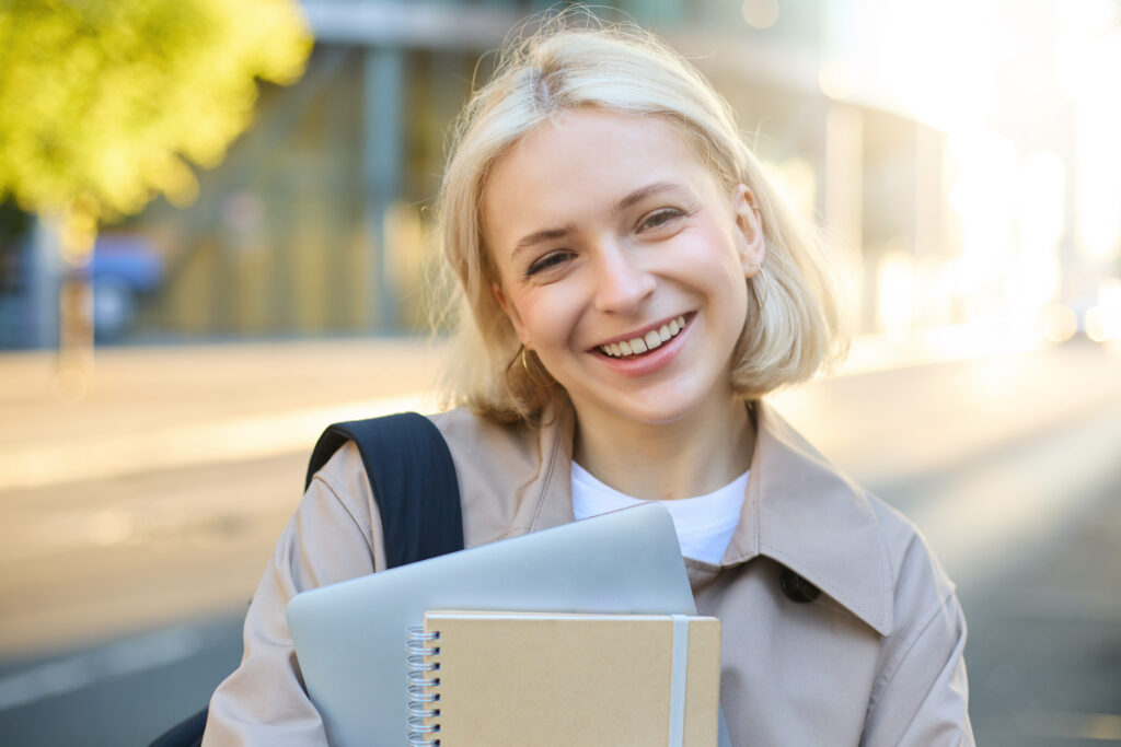 close up portrait smiling blond woman standing street with notebooks carries journal work - „Wspieramy każdego w odkrywaniu i rozwijaniu swojego potencjału, by żyć pełnią życia”